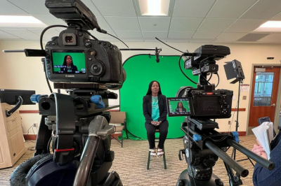 Two video cameras record a woman sitting in front of a green screen.